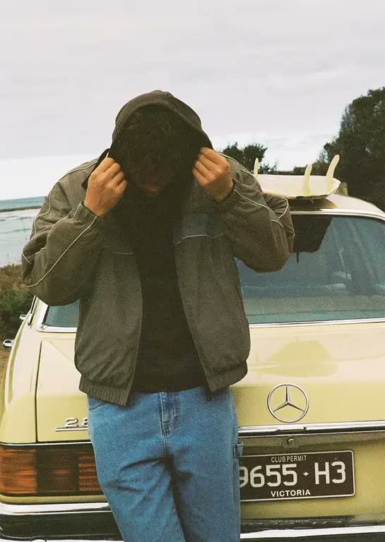 A man in a hooded jacket standing at the back of a cream Mercedes with a surfboard on the roof and Victoria plates