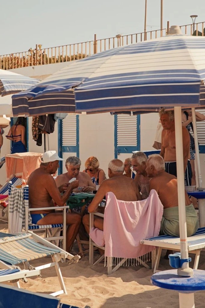 Older men playing cards at a beach club table under blue-and-white striped umbrellas