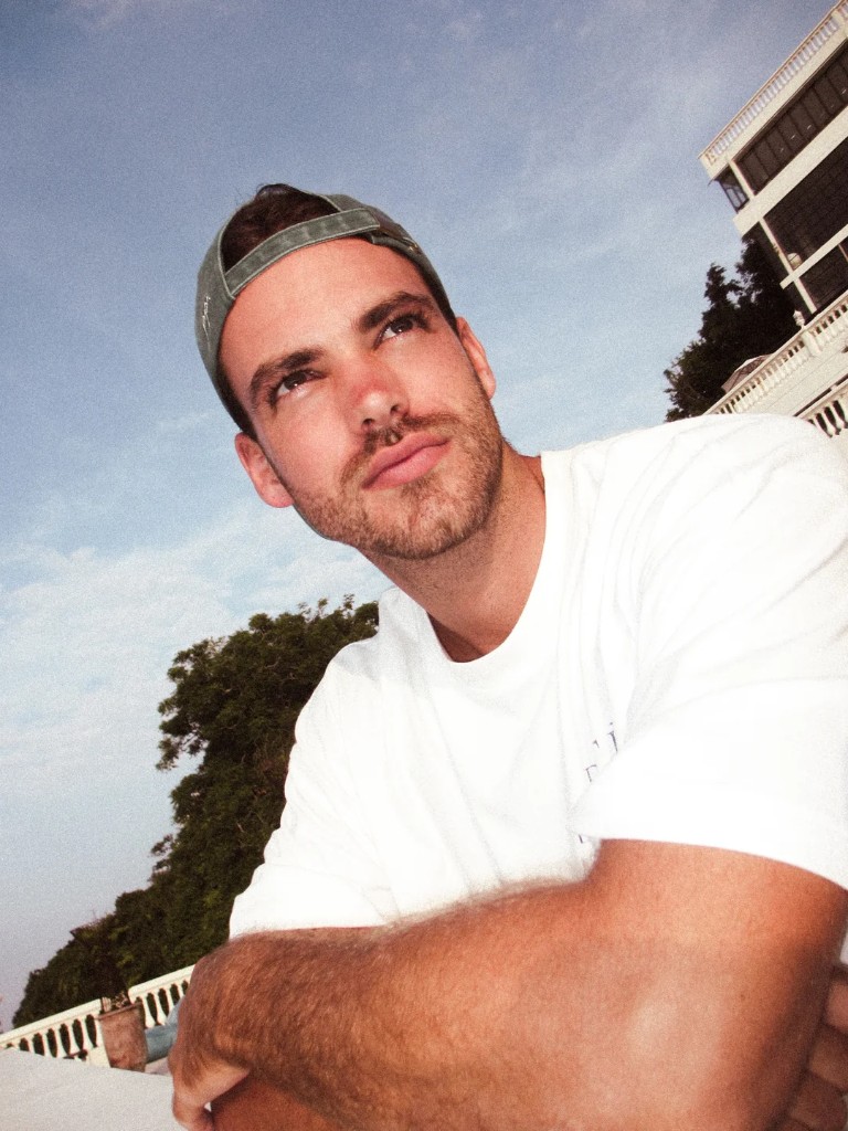 Portrait of a man in a white tee and backwards cap against a coastal sky