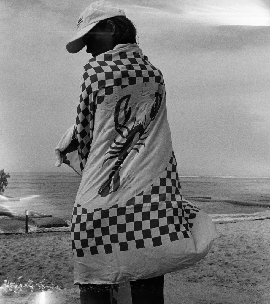Person wrapped in a black-and-white checkered Pete Pareo towel with a lobster print, standing on a beach